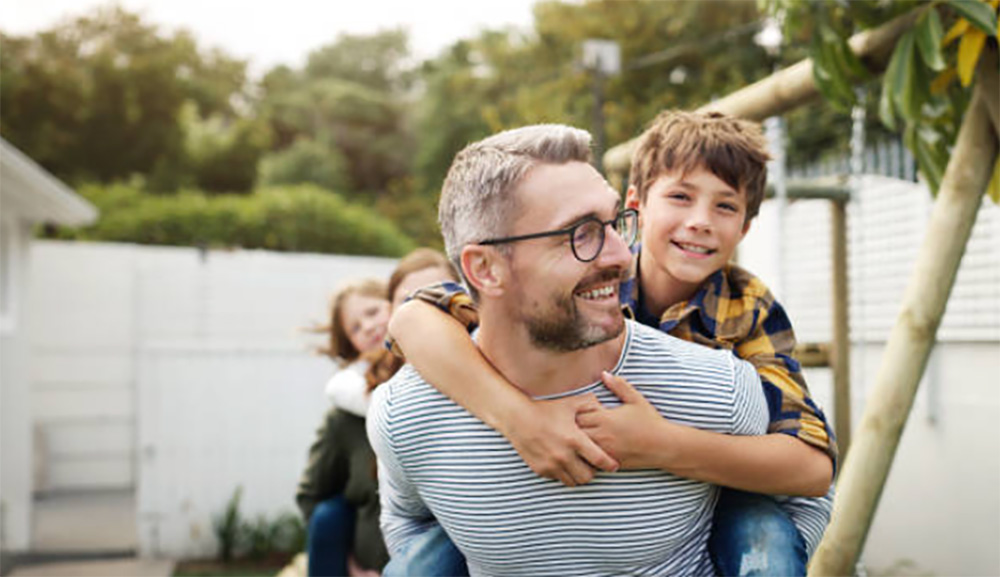 Middle-aged man giving his young son a piggyback ride in their backyard